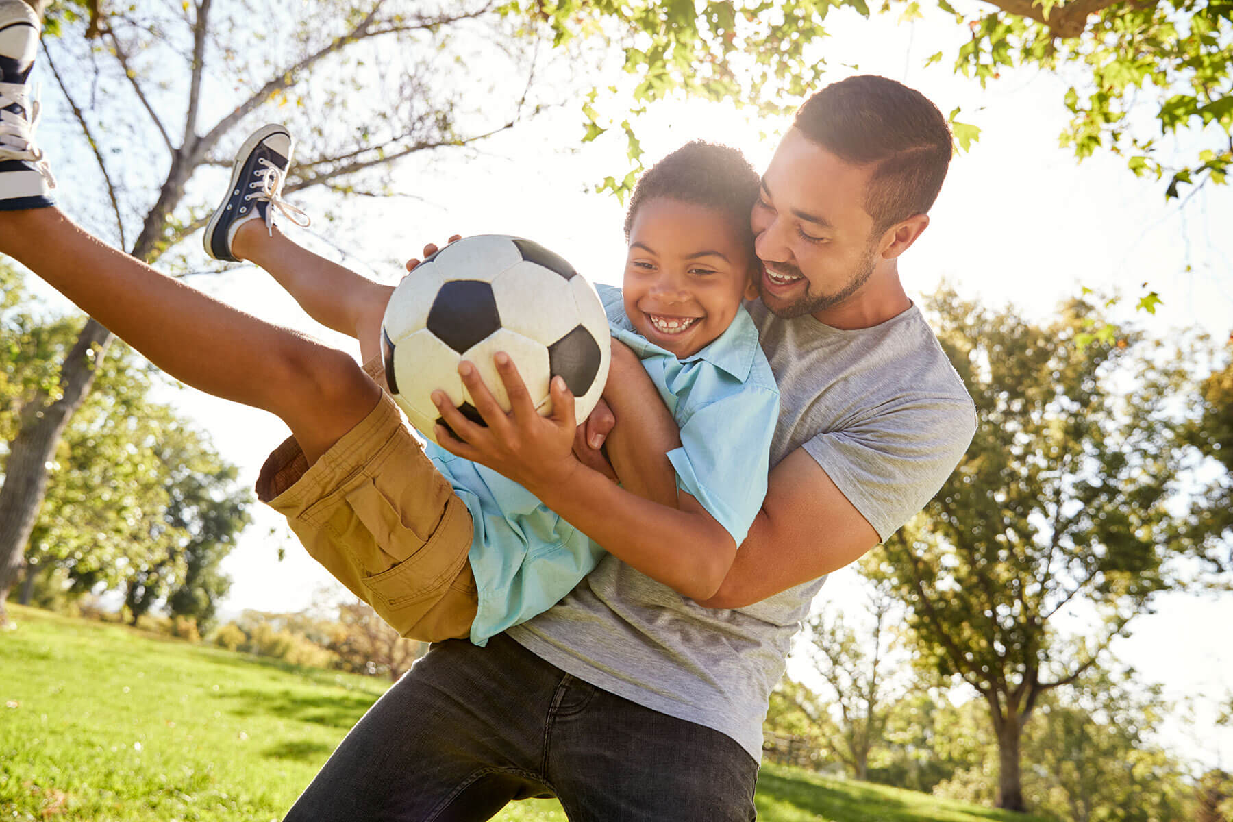 Boy holding a soccer ball getting picked up by his dad in a park